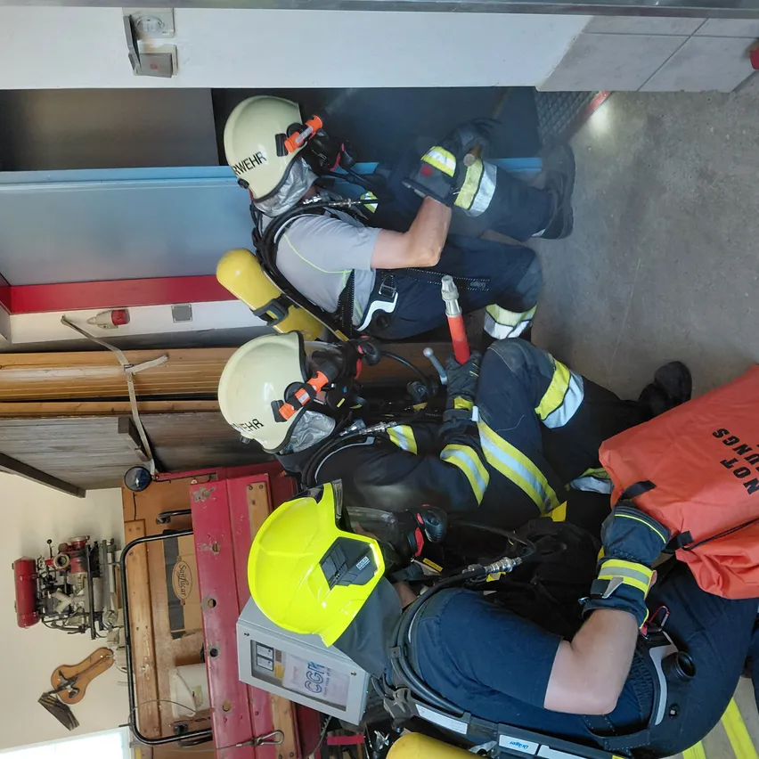 Feuerwehrleute in voller Ausrüstung, einschließlich Helmen und Sauerstoffflaschen, arbeiten in einer Feuerwache. Sie sind in Trainings- oder Rettungseinsätzen tätig.