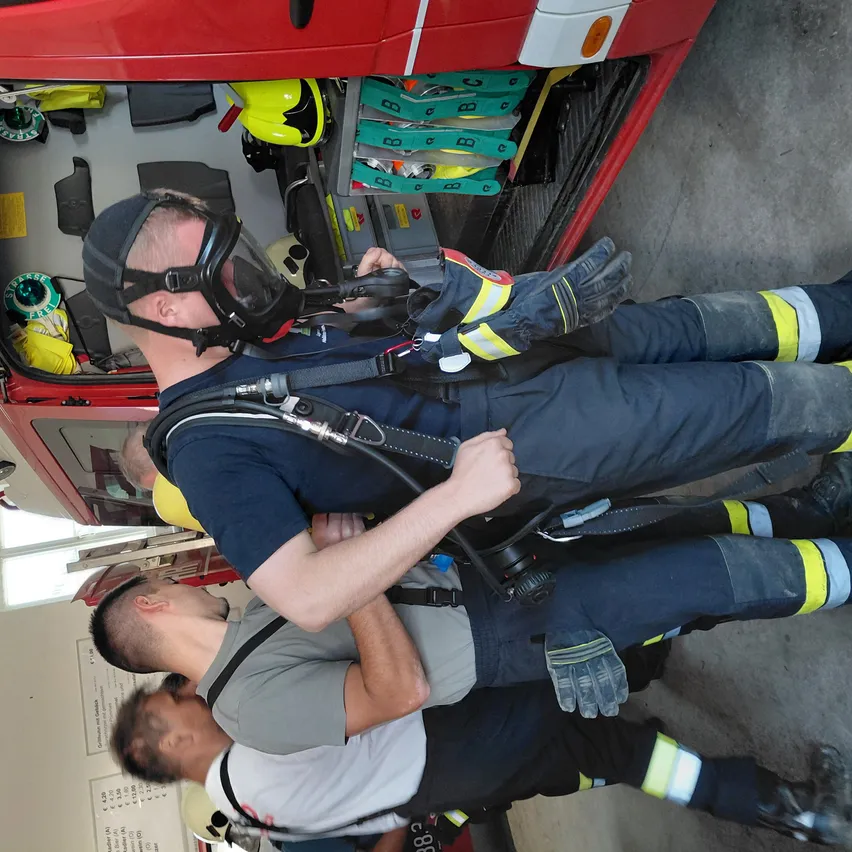Drei Feuerwehrleute in Schutzausrüstung stehen in einem roten Feuerwehrwagen. Einer trägt eine Maske und stützt seine Hand auf die Schulter eines anderen. Sie befinden sich in einer Ausbildungseinrichtung.