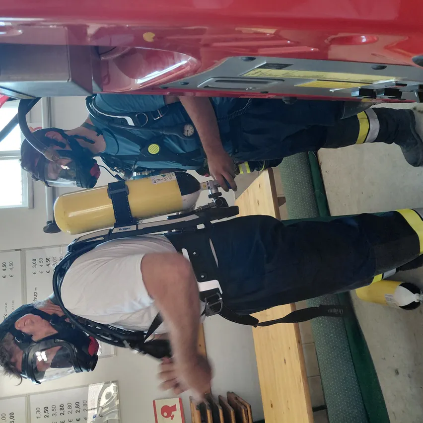 Zwei Feuerwehrleute in voller Ausrüstung führen eine Rettungsaktion in einem roten Fahrzeug durch. Beide tragen Masken und sind mit Sauerstoffflaschen ausgestattet. Der Schauplatz ist in Innenräumen, mit hölzernen Bänken und einer grünen Wand im Hintergrund.