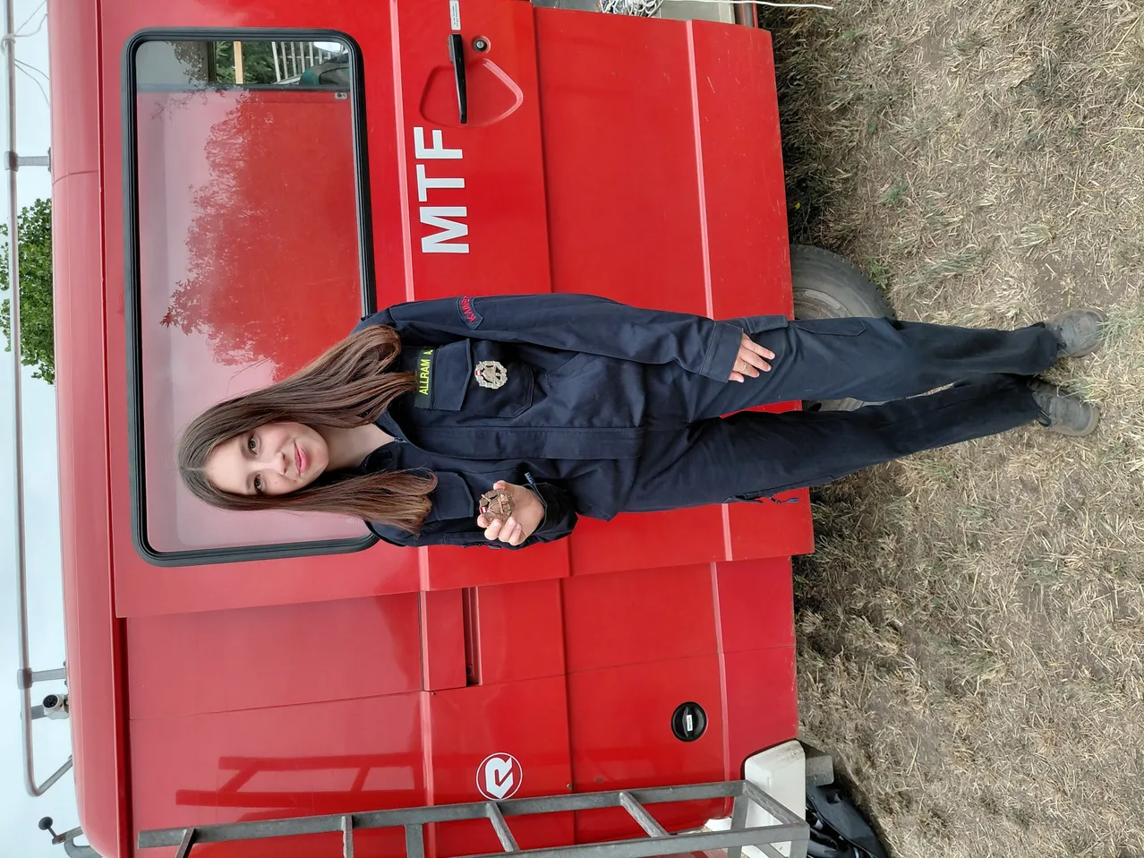 Eine junge Frau in einer blauen Uniform steht neben einem roten Fahrzeug und hält ein kleines Objekt in der Hand. Das Fahrzeug ist mit 'MTf' beschriftet und auf einem Erdboden geparkt.