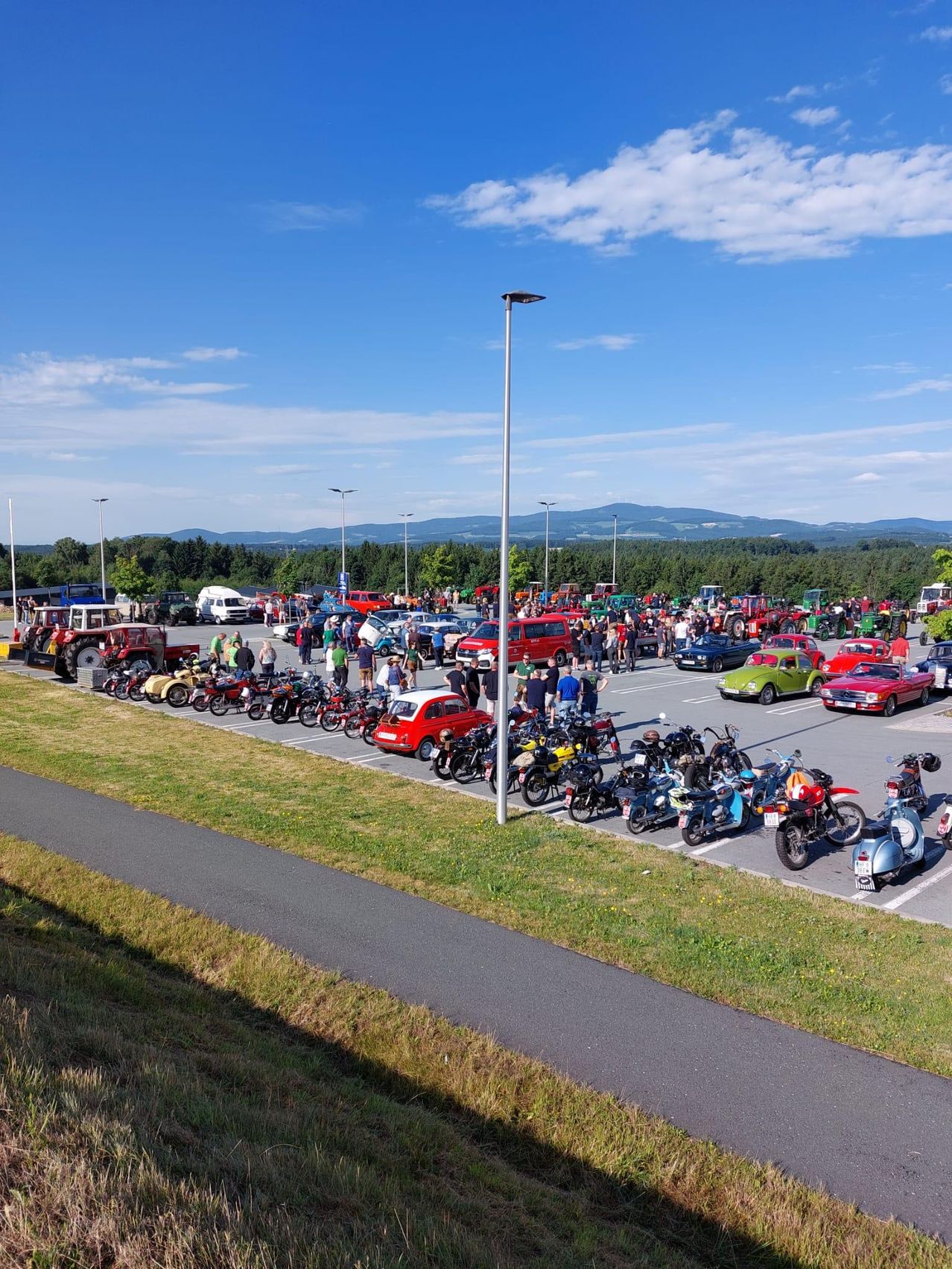 Ein sonniger Außenparkplatz mit Oldtimern, Motorrädern und Traktoren. Viele Menschen sind versammelt, und Berge sind in der Ferne sichtbar.