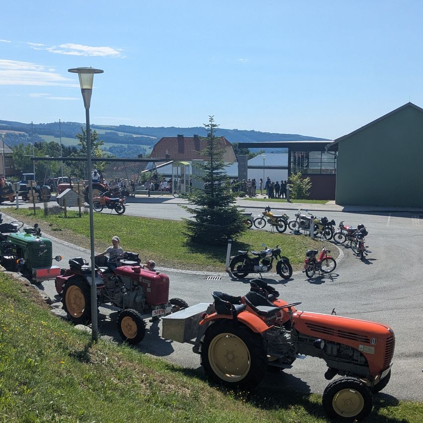Ein sonniger Tag mit Traktoren und Motorrädern, die auf einer Straße geparkt sind, in der Nähe eines Gebäudes und eines Grasbereichs. Menschen stehen im Hintergrund.