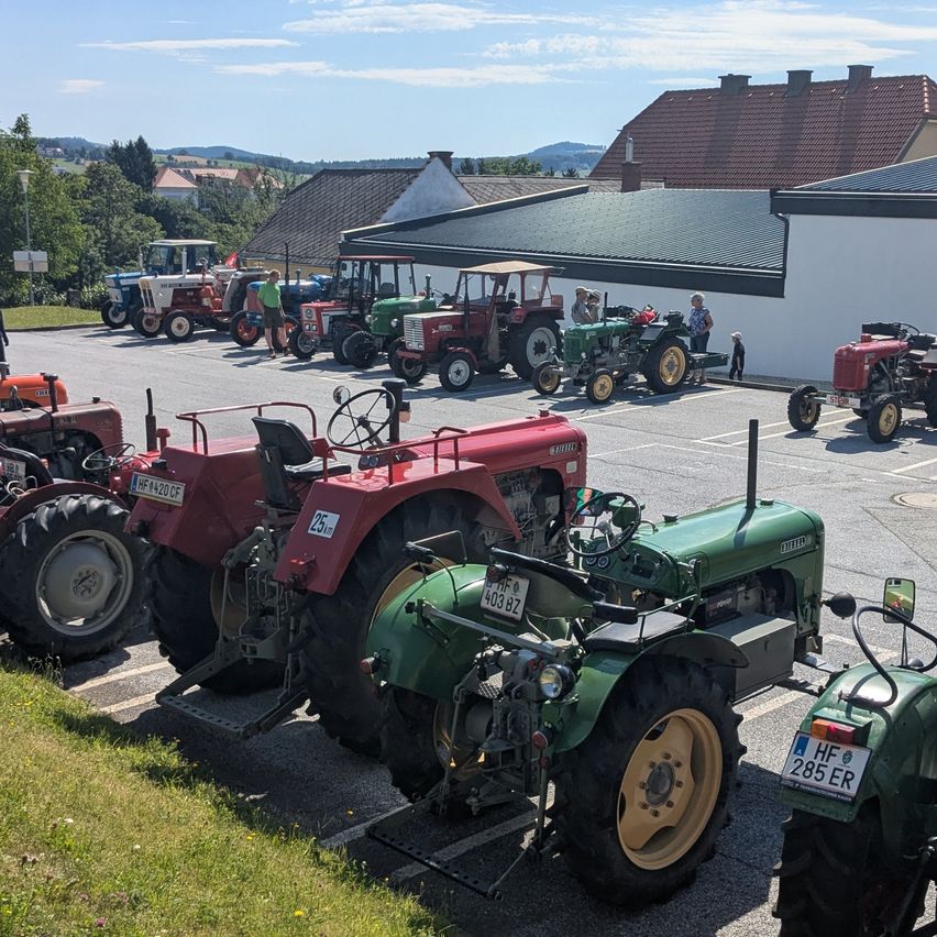 Mehrere Oldtimer-Traktoren sind auf einem Parkplatz abgestellt, mit Menschen, die herumstehen, einer in einem grünen Shirt. Gebäude im Hintergrund, eines mit einem grünen Dach.