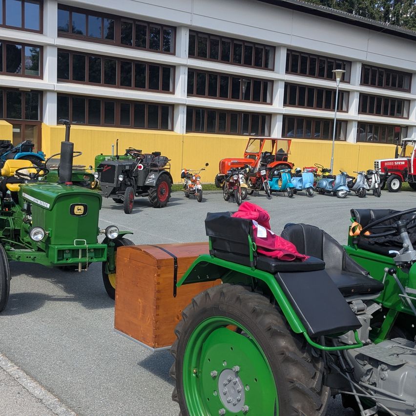 Eine Sammlung von Oldtimer-Traktoren und Rollern ist vor einem Gebäude mit gelben Wänden geparkt. Ein rotes Tuch liegt auf einem der Traktoren.