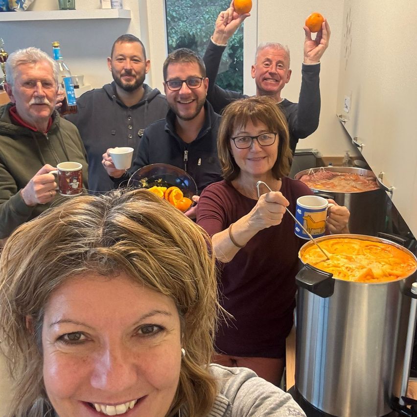 Eine Gruppe von Menschen in einer Küche lächelt und hält Tassen und Orangen. Im Vordergrund steht ein großer Topf mit Essen.