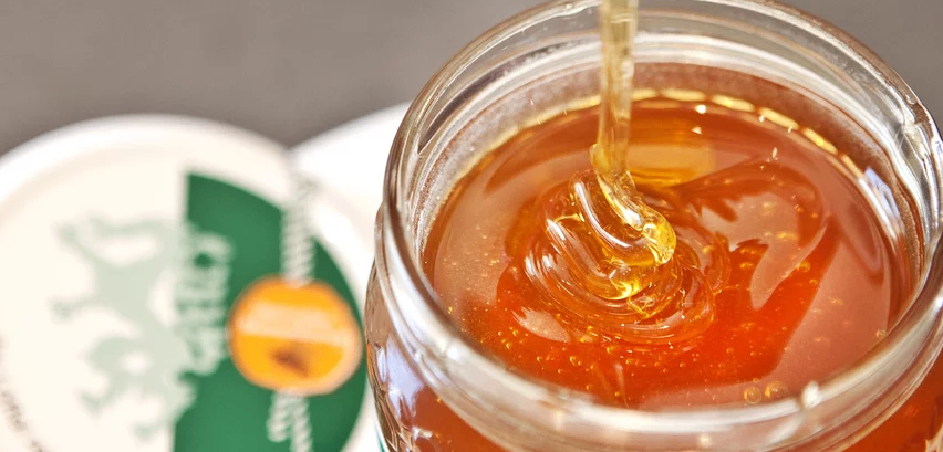 Ein Nahaufnahme eines Glases gefüllt mit bernsteinfarbenem Honig, mit einem goldenen Tropfen, der in das Glas fällt, ein weißer Deckel teilweise im Hintergrund sichtbar.