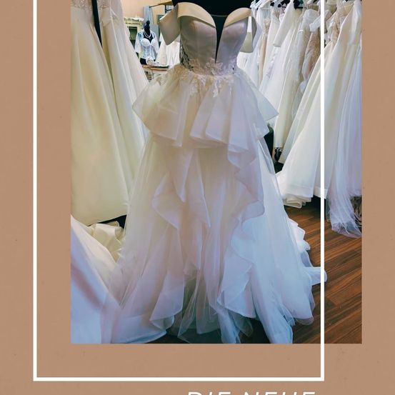Ein weißes Hochzeitskleid mit Rüschen und Spitze wird auf einem Mannequin in einem Brautgeschäft ausgestellt. Das Kleid ist von anderen Hochzeitskleidern umgeben.