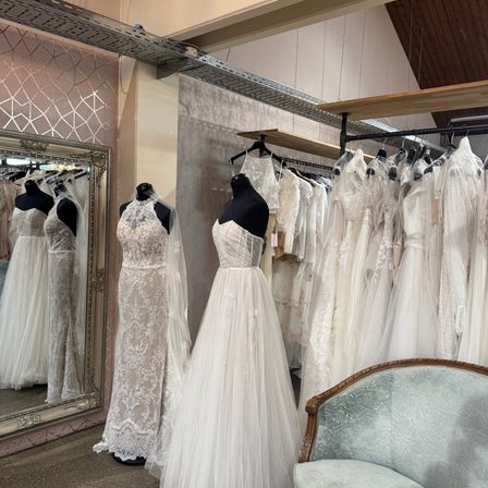 Ein Brautmodengeschäft mit einer Vielzahl von Brautkleidern, die auf Mannequins und Gestellen ausgestellt sind, mit einem Spiegel, der die Kleider reflektiert, und einem Stuhl im Vordergrund.