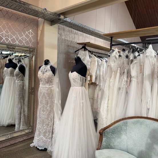 Ein Brautmodengeschäft mit einer Vielzahl von Brautkleidern, die auf Mannequins und Gestellen ausgestellt sind, mit einem Spiegel, der die Kleider reflektiert, und einem Stuhl im Vordergrund.