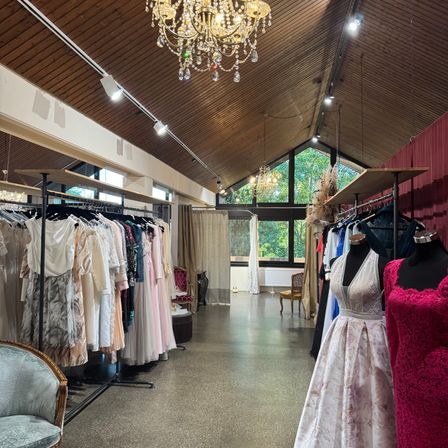 Ein Innenansicht eines Boutiques, das eine Vielzahl von Kleidern auf Gestellen präsentiert, mit Kronleuchtern und natürlichem Licht durch Fenster. Mannequins zeigen Kleider im Vordergrund.