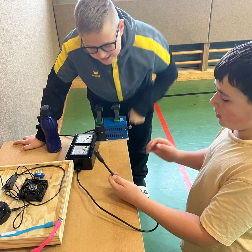 Zwei Jungen arbeiten in einem Klassenzimmer an einem Wissenschaftsprojekt. Einer steht und der andere sitzt. Beide benutzen ein Gerät mit Kabeln.