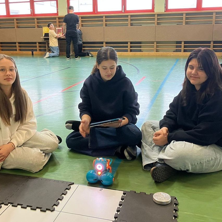 Drei Mädchen sitzen auf dem Boden einer Turnhalle, zwei tragen Kapuzenpullover, eines Brille. Eines hält ein Tablet, ein anderes schaut einen Spielzeugroboter an. Ein Junge kniet an einem Tisch, eine weitere Person steht hinter ihm.