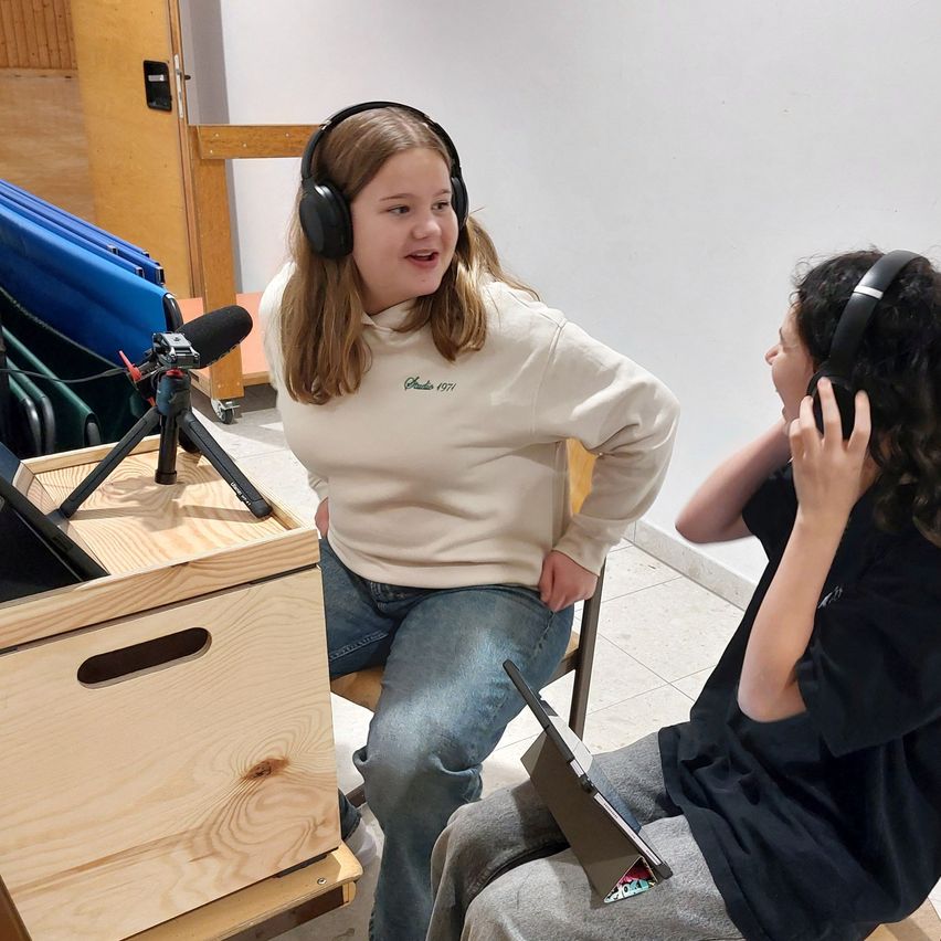 Zwei junge Menschen sitzen in einem Aufnahmestudio. Ein Mädchen mit Kopfhörern lächelt, während ein Junge neben ihr aufmerksam zuhört. Sie haben ein Mikrofon und eine Holzkiste auf dem Boden.