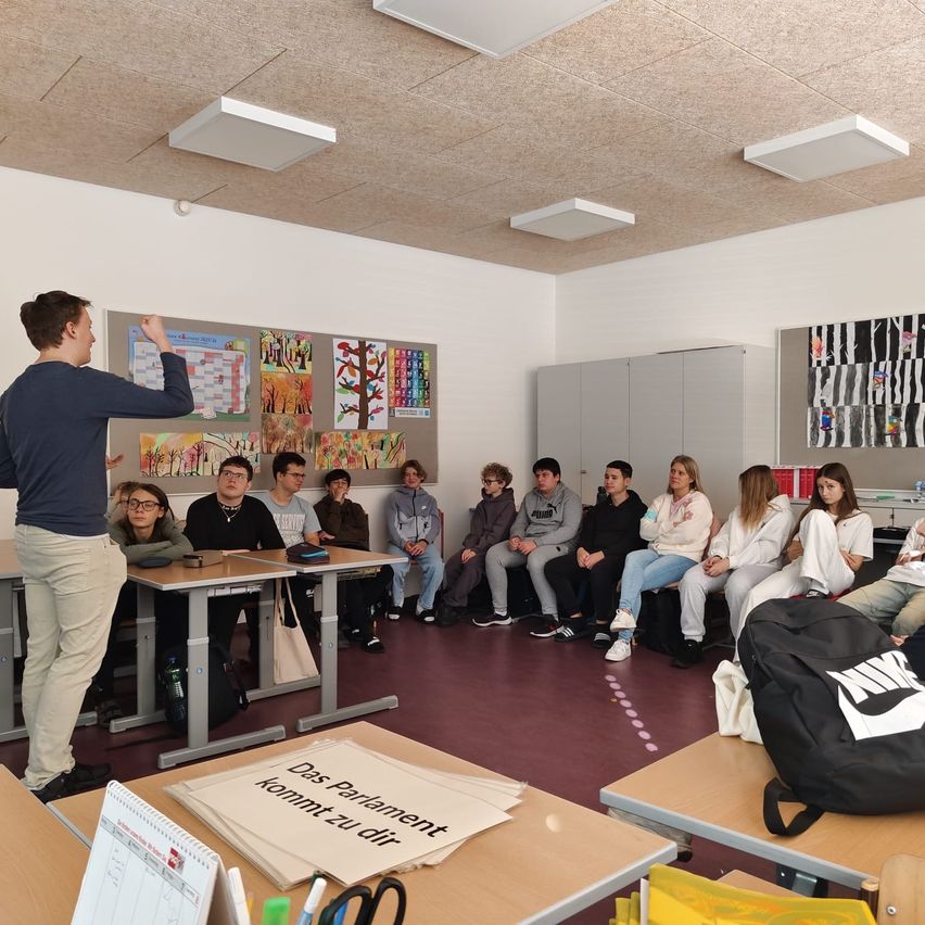 A classroom with students sitting at desks. A man stands in front of a board. Art posters are on the wall. A backpack is on a desk.