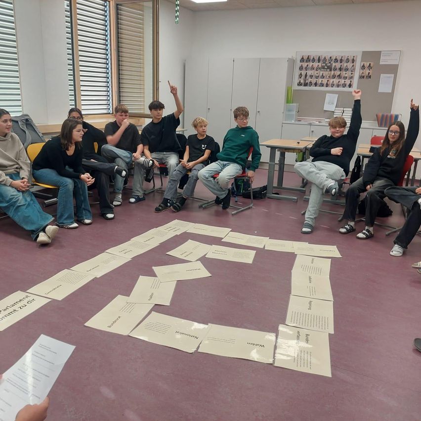 Students are sitting in a circle on the floor, forming a maze with papers. A few students have their hands raised. The room has desks, cabinets, and bulletin boards.