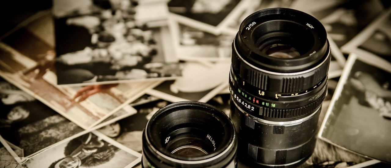 Zwei Vintage-Schwarzkameraobjektive liegen auf einem Stapel alter Fotos mit Menschen und unscharfen Bildern.