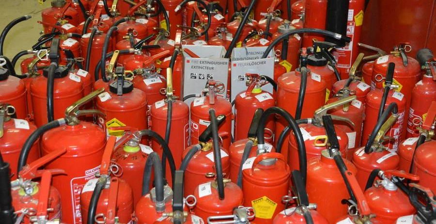 Ein Raum gefüllt mit zahlreichen roten Feuerlöschern, jeder mit weißen Texten beschriftet und mit schwarzen Griffen und Schläuchen ausgestattet. Ein Schild im Hintergrund weist 'Feuerlöscher' in mehreren Sprachen aus.