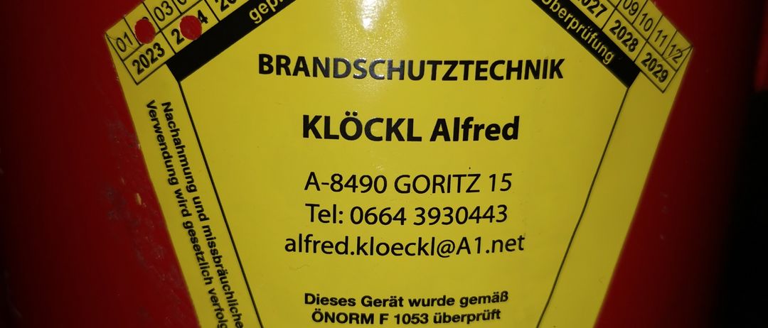 Ein gelber Aufkleber mit sechseckiger Form. Er zeigt den Firmennamen 'Brandschutztechnik Klockl Alfred', die Adresse 'A-8490 Goritz 15', Kontaktinformationen und eine TUV Austria Zertifizierung. Der Aufkleber enthält auch einen Wartungsplan.