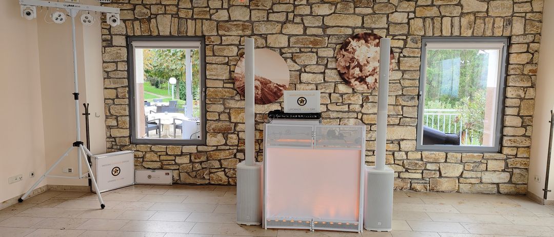 Ein Raum mit einer Steinmauer, einer Glastür und einem Lautsprecher-Setup mit Laptop. Es gibt zwei runde Wanddekorationen mit Blumen und einem rosa Licht.