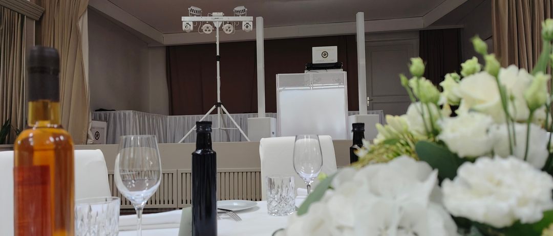 Ein Tisch mit weißen Blumen, Weingläsern und einer Flasche, die für eine Veranstaltung vorbereitet ist. Im Hintergrund steht ein Podest mit Laptop, Mikrofon und Beleuchtungsequipment.