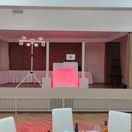Eine leere Bühneneinrichtung mit pinken Lichtern auf einem Ständer, einer DJ-Box mit Laptop und einem weißen Vorhang im Hintergrund. Im Vordergrund sind Stühle mit Weingläsern und Flaschen auf einem Tisch angeordnet.