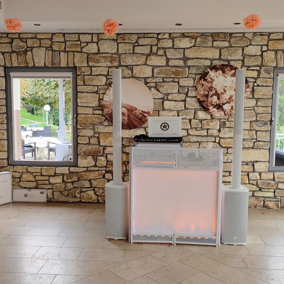 Ein Raum mit einer Steinmauer, der zwei große Lautsprecher, eine beleuchtete Konsole und zwei kreisförmige Blumendekorationen an der Decke aufweist. Vor dem Fenster befindet sich ein Garten mit Bäumen und einem Tisch mit Stühlen.