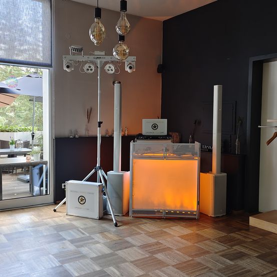 Ein Innenbereich zeigt eine Beleuchtungseinrichtung mit mehreren Lampen, einer Kiste auf einem Stativ und einem glasüberdachten Schreibtisch. Der Raum hat Holzböden, eine Glastür und einen Tisch mit Pflanzen.