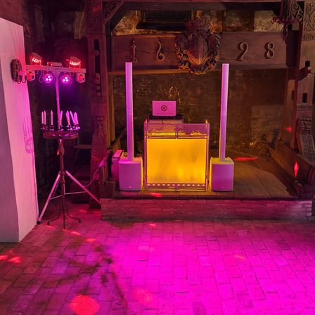 Eine Bühne mit DJ-Setup, bestehend aus einer gelben Lichtbox, Lautsprechern und Kerzen auf einem Ständer. Der Raum hat eine Vintage-Atmosphäre mit Holzwänden und einem Ziegelboden.