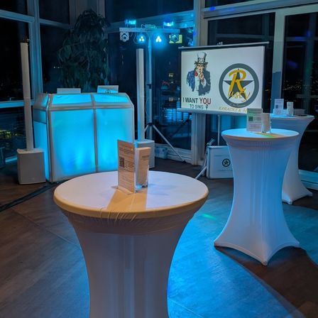 Ein Raum mit einer Karaoke-Einrichtung, mit einem großen Bildschirm mit einem Poster von Uncle Sam und einem Banner. Zwei weiße runde Tische mit Gegenständen darauf und einem großen beleuchteten Lautsprecher.