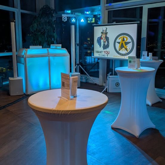 Ein Raum mit einer Karaoke-Einrichtung, mit einem großen Bildschirm mit einem Poster von Uncle Sam und einem Banner. Zwei weiße runde Tische mit Gegenständen darauf und einem großen beleuchteten Lautsprecher.