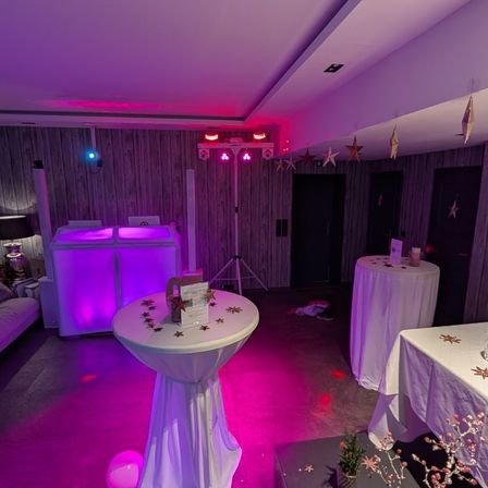Ein Raum mit DJ-Pult, Tischen und lila Beleuchtung. Das DJ-Pult hat eine lila LED-Leuchte oben. Die Tische sind mit weißen Tüchern bedeckt. Der Raum hat eine Holzwand und eine Decke mit Sternen, die daran hängen.