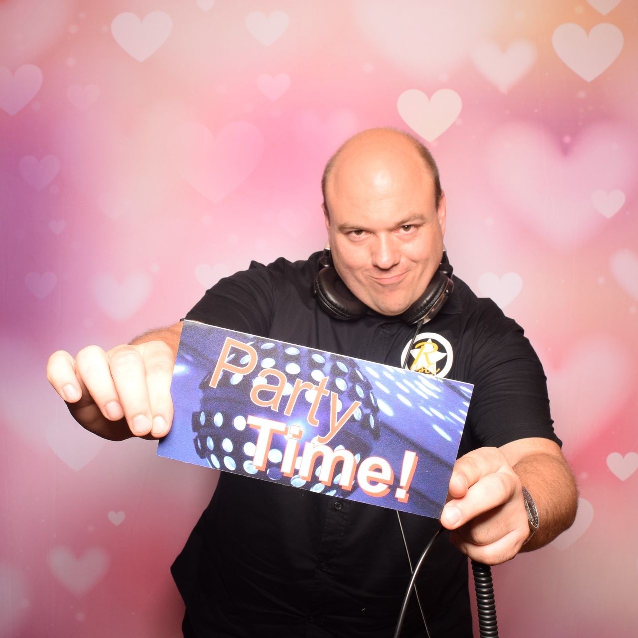 Ein Mann mit Kopfhörern hält ein Schild, auf dem 'Party Time!' steht. Er lächelt und posiert für ein Foto. Der Hintergrund ist pink mit Herzformen.