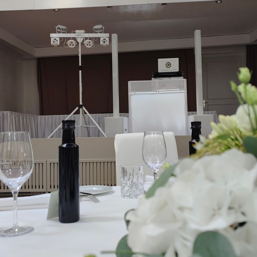 Ein Essplatz mit weißer Tischdecke, weißen Blumen, zwei Weingläsern und einer Flasche. Im Hintergrund steht ein Rednerpult mit Lautsprecher und Laptop.