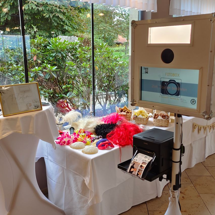 Ein Fotoautomat mit verschiedenen Requisiten wie Hüten und Blumen ist in einem Raum mit Glaswänden aufgestellt, der einen Blick auf einen Garten bietet.