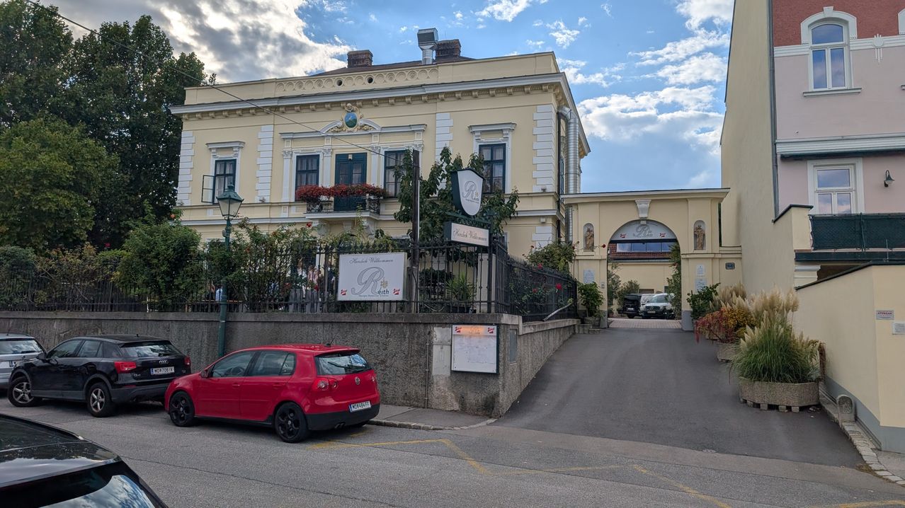 A red car is parked on the side of the road in front of a building with a sign that reads 'Ruth'. The building has a balcony with potted plants and a black fence.