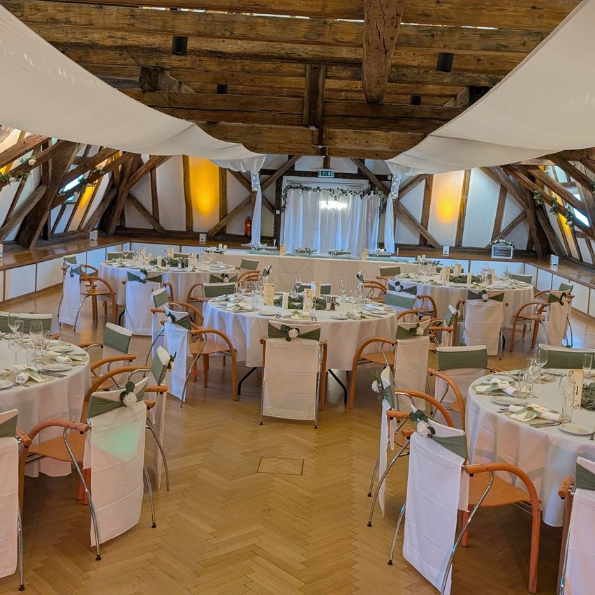 Ein Esssaal mit Holzdecke und weißen Tüchern, die darüber hängen. Runde Tische mit weißen Tüchern und Dekorationen sind mit Stühlen aufgestellt. Es gibt eine Bühne mit einem weißen Vorhang im Hintergrund.