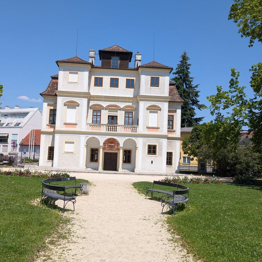 Ein weißes Gebäude mit braunen Akzenten, mehreren Fenstern und einer großen Holztür steht in einem Park. Das Gebiet ist von grünem Rasen und Bäumen umgeben. Zwei Bänke befinden sich auf dem Weg zum Gebäude.
