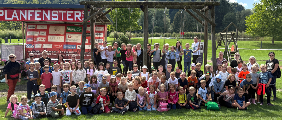Eine große Gruppe von Kindern, meist in weißen Hemden, posiert für ein Foto vor einer hölzernen Schaukel. Im Hintergrund befindet sich ein rotes Gebäude und Bäume.