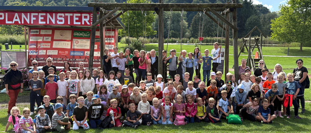 Eine Gruppe von Kindern und Erwachsenen posiert für ein Foto unter einer Schaukelkonstruktion in einem Park mit grünen Bäumen und einem Gebäude im Hintergrund.