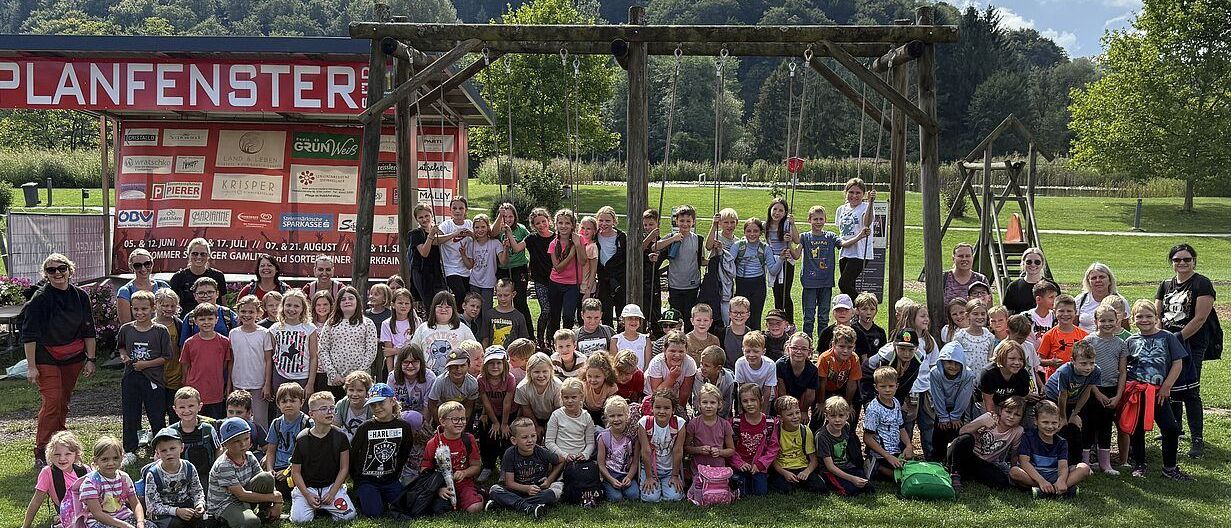 Eine Gruppe von Kindern und Erwachsenen posiert für ein Foto auf einem Rasenfeld mit einer Holzschaukel im Hintergrund. Bäume und Berge sind in der Ferne zu sehen.