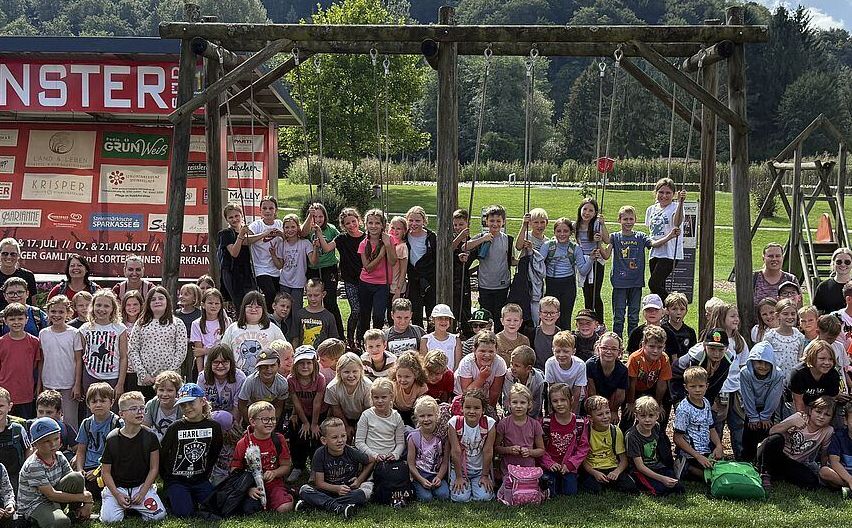 Eine Gruppe von Kindern und Erwachsenen posiert für ein Foto auf einem Rasenfeld mit einer Holzschaukel im Hintergrund. Bäume und Berge sind in der Ferne zu sehen.