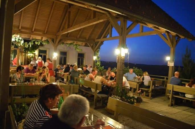 Ein Outdoor-Restaurant mit einem hölzernen Pavillon ist gefüllt mit Menschen, die bei Nacht essen, unter warmen Lichtern und klarem Himmel.