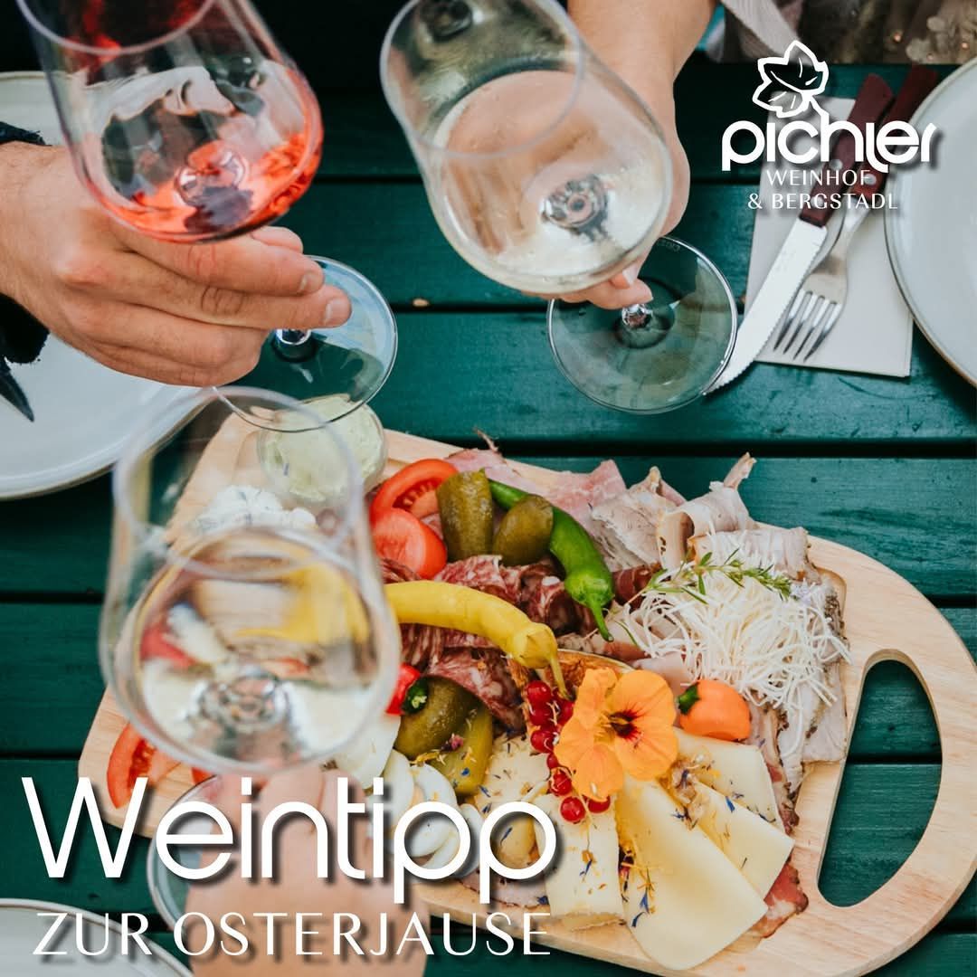 Zwei Hände stoßen mit Weingläsern über einem Holzbrett mit Essen und Blumen an. Das Brett steht auf einem grünen Tisch mit Tellern, Besteck und einer weißen Serviette.