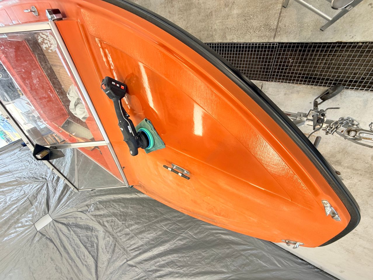 Ein orangefarbenes Boot mit schwarzem Griff und grünem Tuch wird mit einem Werkzeug poliert. Das Boot hat eine silberne Umrandung und liegt auf einem grauen Tuch.