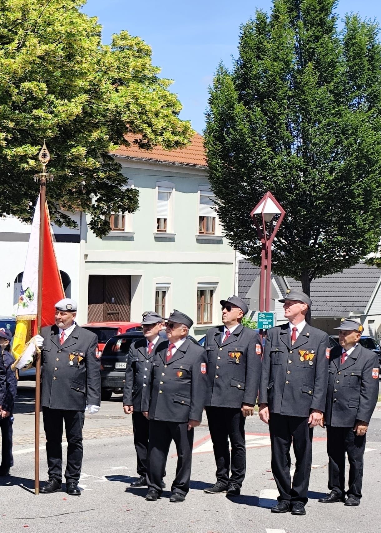 A group of uniformed men are standing in a row, holding a flag in the front. They are on the side of the road, with a building, cars, and trees in the background.