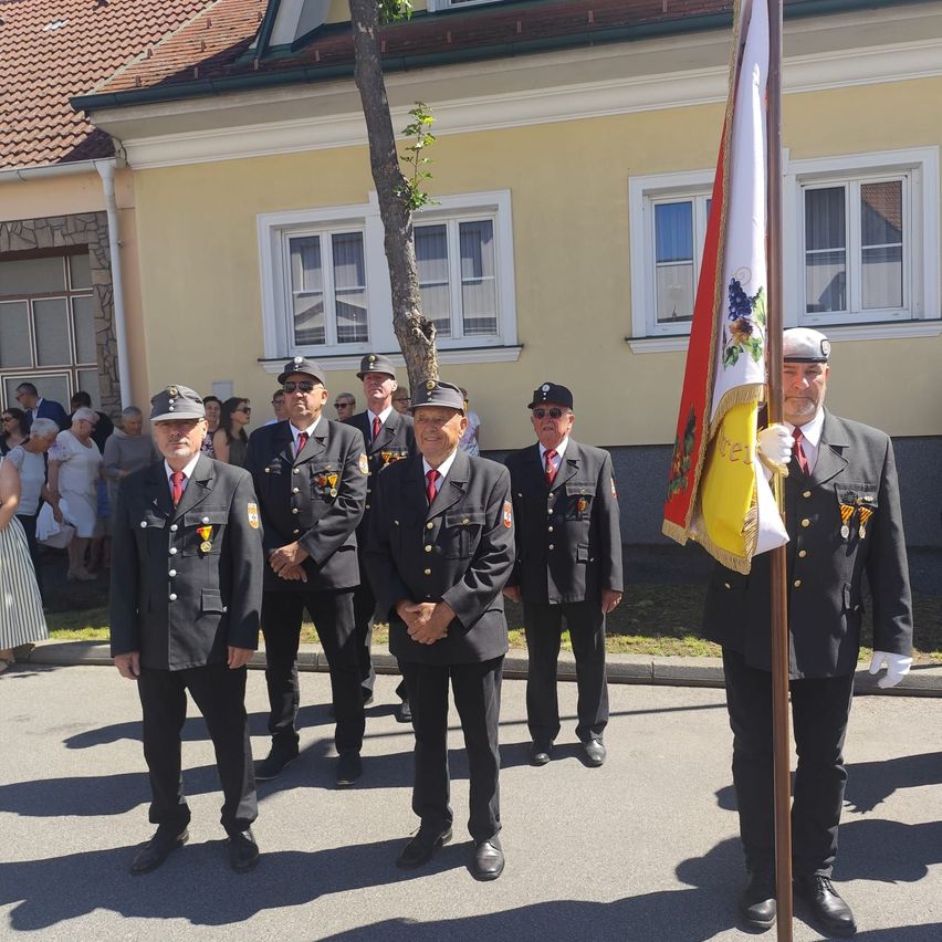 Six men in uniform, holding flags, stand in front of a building. Spectators are in the background.