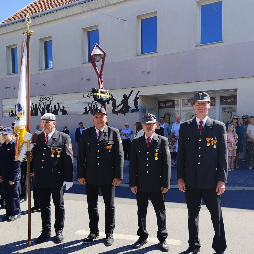 Four men in uniform stand in front of a building with a banner. They are likely part of a ceremonial event. Behind them are several people observing. The building has windows and a sign that reads 'DISCO'.