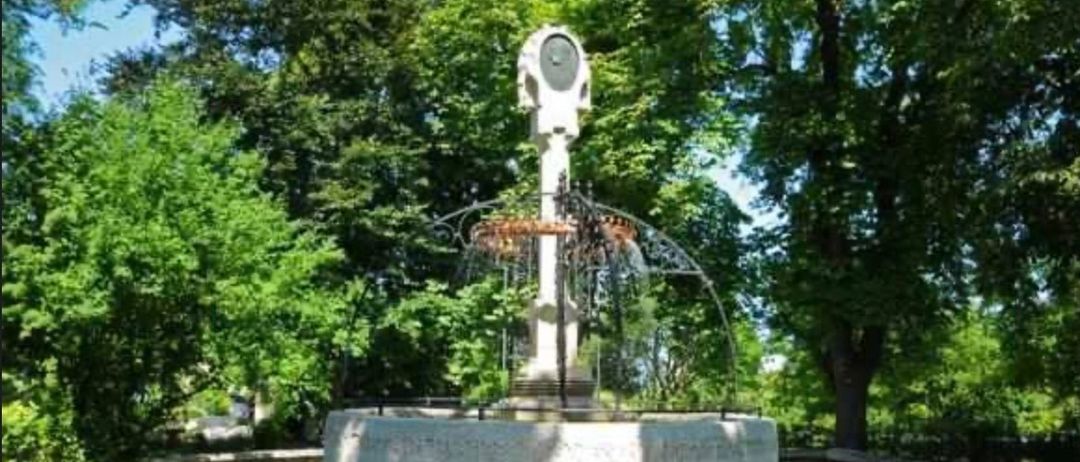 Ein Steinbrunnen mit einer weißen Statue ist von einem Metallzaun umgeben. Es befindet sich in einem Park mit Bäumen.