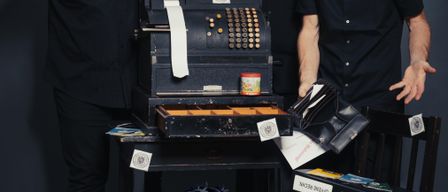 Drei Männer stehen um eine Kasse herum, halten einen Hammer, Sparschwein und eine Mappe. Die Kasse hat Papiere und Ordner auf einem Tisch. Termine 14., 15. und 16. Februar 2026. Eintritt 12 Euro, Kinder 9 Euro.