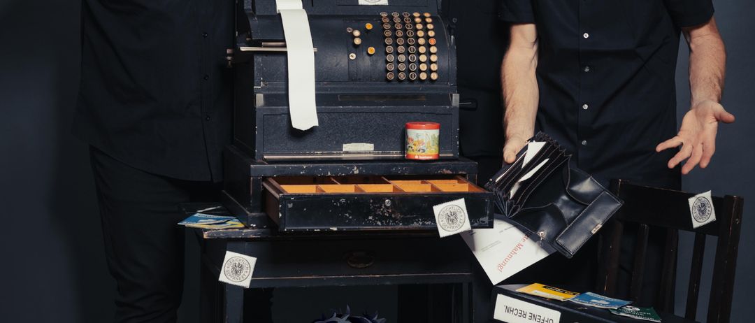 Drei Männer stehen um eine Kasse herum, halten einen Hammer, Sparschwein und eine Mappe. Die Kasse hat Papiere und Ordner auf einem Tisch. Termine 14., 15. und 16. Februar 2026. Eintritt 12 Euro, Kinder 9 Euro.